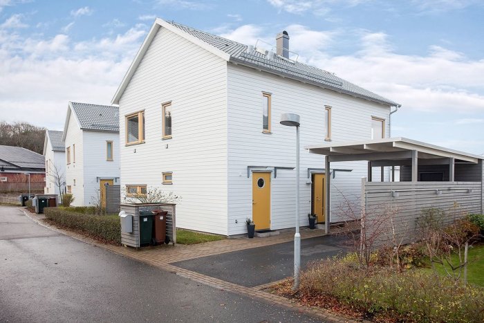 Vit panelhus med gult dörr, carport, stenlagd uppfart och soptunnor vid sidan.