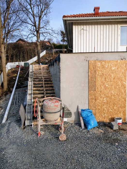 En nygjuten trappa i betong som leder upp till en gäststuga med byggmaterial och en cementblandare i förgrunden.