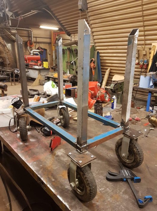 Hjulförsedd metallvagn under konstruktion med fyra stöttor och breddningsplattor i en verkstadsmiljö.
