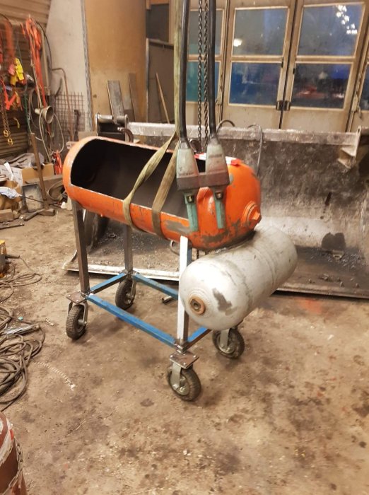 En orange betongblandare på en blå vagn, upphängd med spännband, med en gasoltub vid sidan i en verkstad.