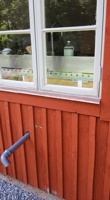 Nymålad röd stuga med vitt fönster och grått dräneringsrör längs väggen.