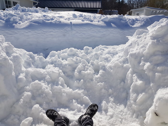 Person som sitter i djup snö med fötterna framtill, snötäckta hus och blå himmel i bakgrunden.