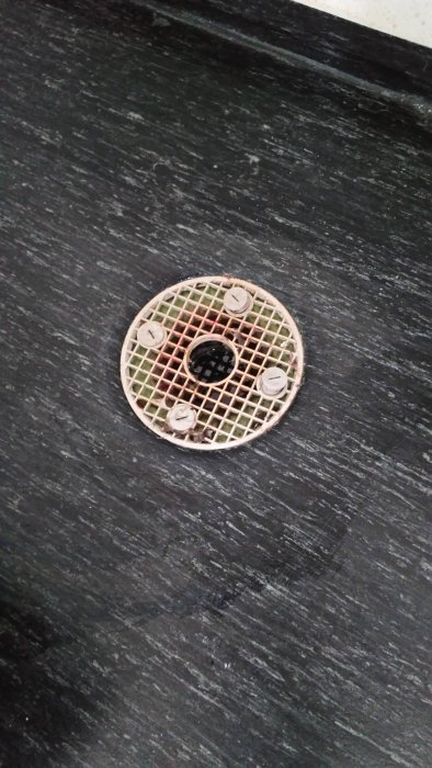 Golvbrunn med metallgaller på en mörk kaklad yta, skruvar synliga, vissa områden verkade rensade.