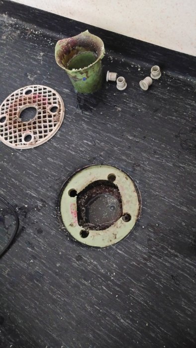 Demonterad golvbrunn med borttaget galler, synlig klämring och samlad smuts på mörkt golv.