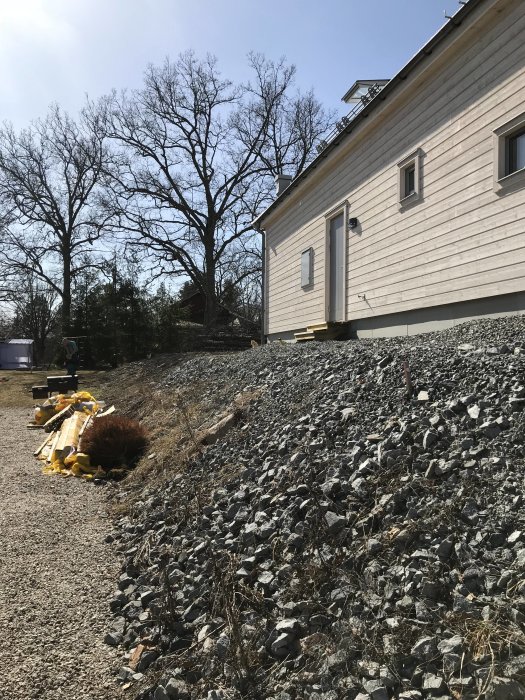 Brant slänt med stenar intill en husvägg som indikerar behov av stödmur för att förhindra erosion och säkerställa markstabilitet.