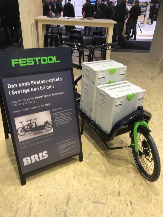 Grön-svart lastcykel framför en skylt med texten "FESTOOL" och lådor vid en mässa.