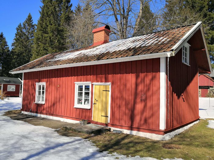 Traditionellt rött trätorp med vita knutar och gult dörr, snötäckt tak och smältande snö på marken.