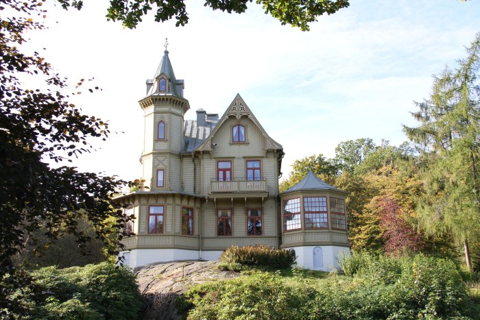 Traditionellt trähus i 1890-talsstil med torn, snickarglädje, utsmyckade verandor och omgivet av träd.