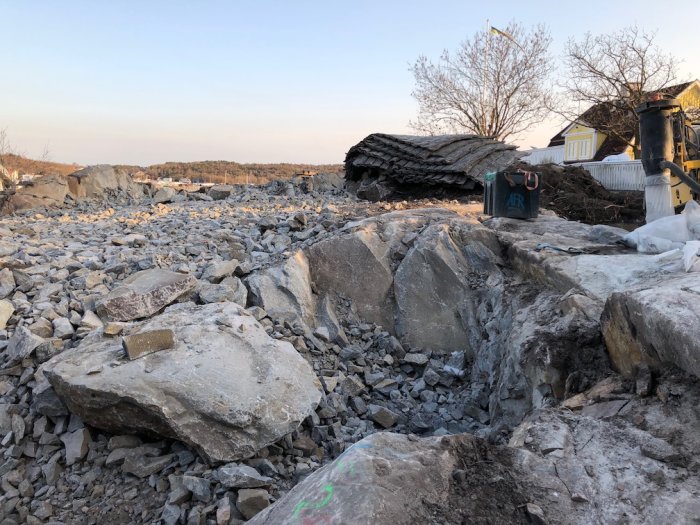 Schaktbotten med stora stenblock och grus vid ett byggprojekt, sprängningsarbete pågår, med byggnad i bakgrunden.