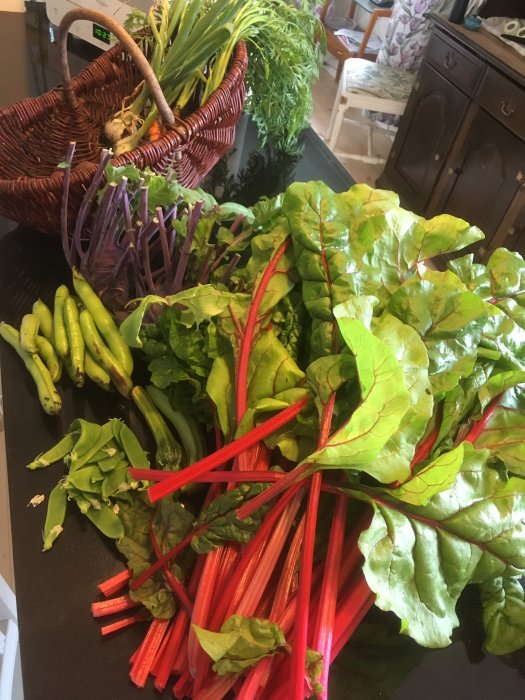 Färska trädgårdsgrönsaker inklusive rödbetor, ärtor och bladgrönt på ett köksbord med en korg i bakgrunden.