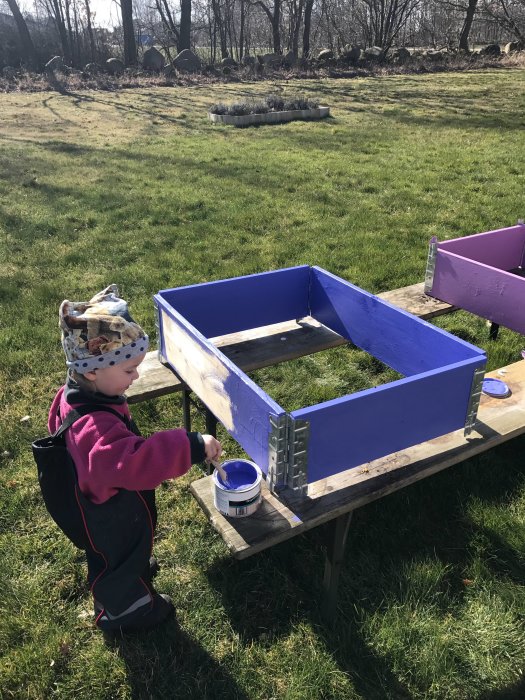 Barn målar en hemmagjord lila odlingslåda utomhus på en solig dag.