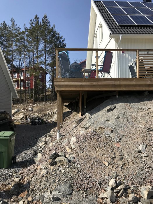 Trädäck med räcke framför ett hus, omgivet av skog och solpaneler på taket.