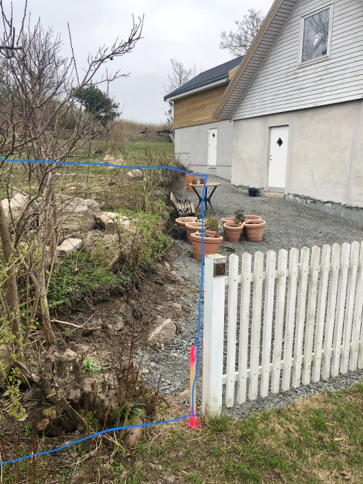 Trädgårdsutsikt av bakre huset med sluttande tak, grusgång och ett vitt staket i förgrunden, med krukor och grönska.