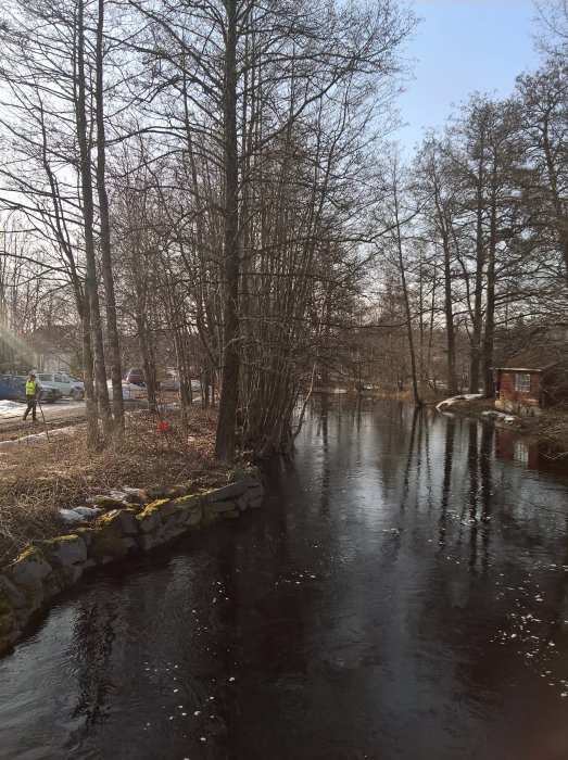 Person joggar på en byväg bredvid en isbelagd å med träd och lite snö på marken.