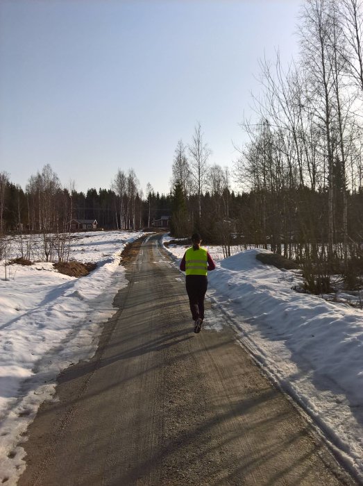 Joggare i reflexväst på vårbefriad grusväg omgiven av snötäckta fält och skog.