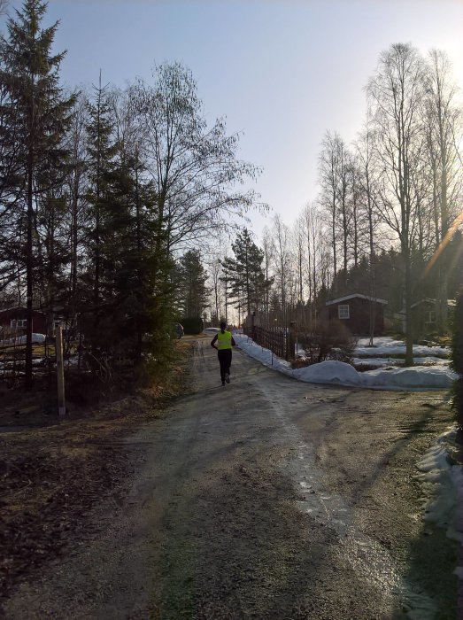 Person i löpkläder joggar på en grusväg i bymiljö med snökanter och träd i bakgrunden.