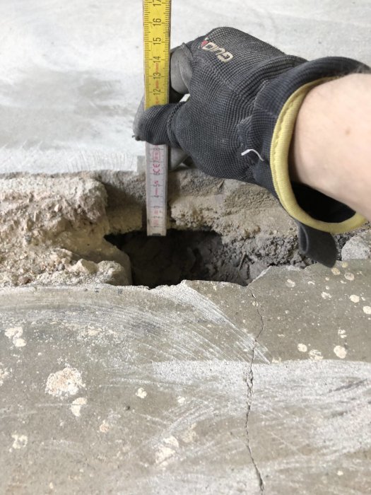 Hand med skyddshandske mäter ett hål i en slitad betongplatta med sprickor och limrester, med tumstocken synlig.