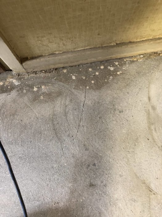 Spricka i betonggolv nära vägg med synliga rester av lim och gamla trösklar.