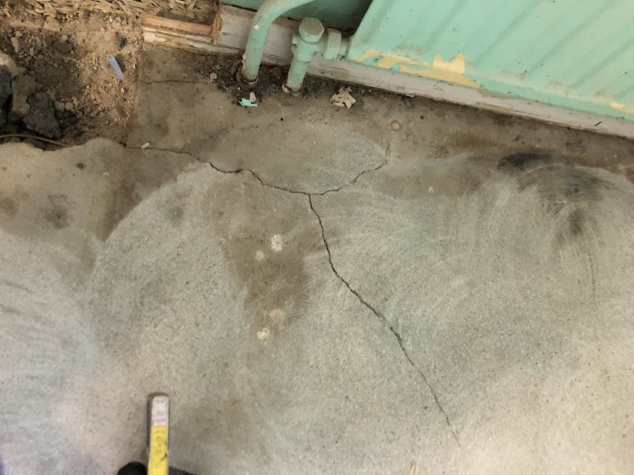 En konkret platta med sprickor och hål nära vattenrör och en gammal väggs position, mätverktyg synligt.