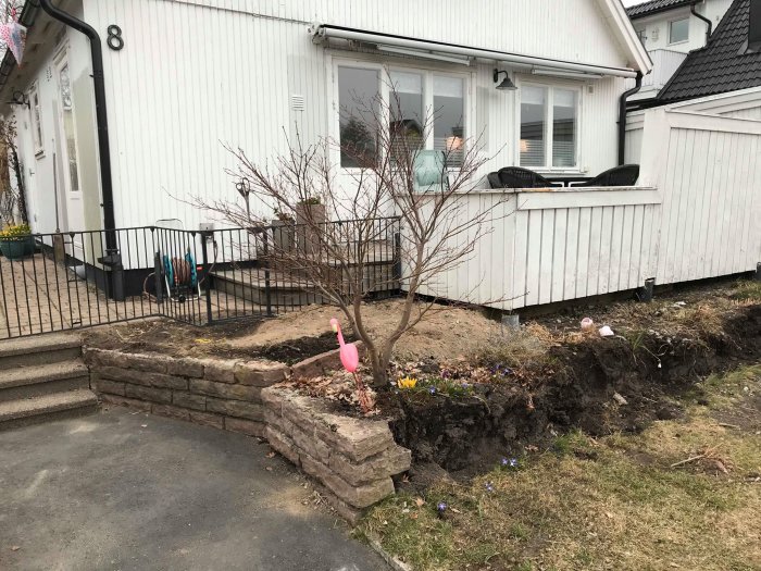 Delvis riven stödmur framför vit husfasad med gammal altan och en bar trädstam, omringad av jord och sparsam vegetation.