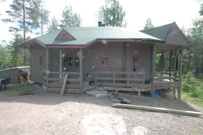 Ett grått timmerhus med rosa fönsteromramningar och en veranda under en klar himmel, omgivet av skog.