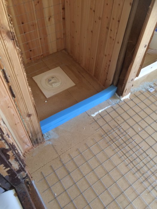 Hörn av ett badrum under renovering med synligt golvavlopp, blå tejpning och halvfärdigt kakelarbete.
