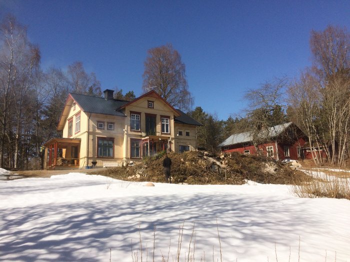 Stort gult hus under renovation med trädstammar på marken, omgivet av snö och en röd lada i bakgrunden.