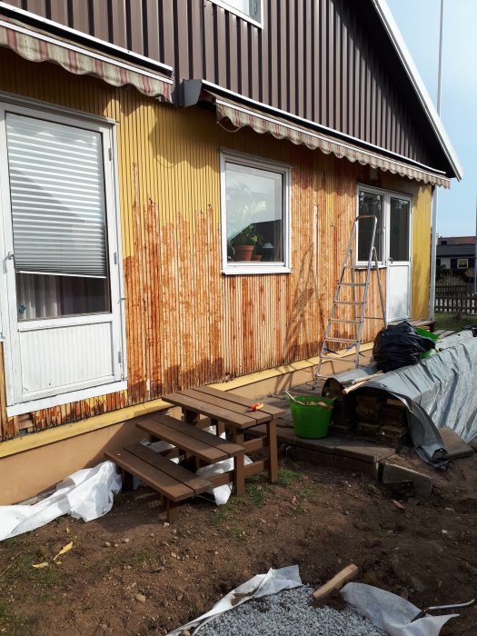 Hus med delvis avlägsnat yttre aluminiumskikt på panelen, vit ärg synlig på fasaden, byggmaterial och trappa i förgrunden.