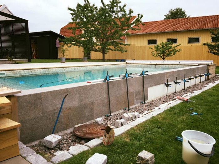 Pool med frigolitvägg där keramiska plattor är under uppsättning, omgiven av trädgårdsverktyg och renoveringsmaterial.