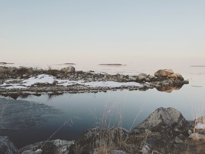 Vinterlandskap vid havet med stenar och snö under en klar himmel.