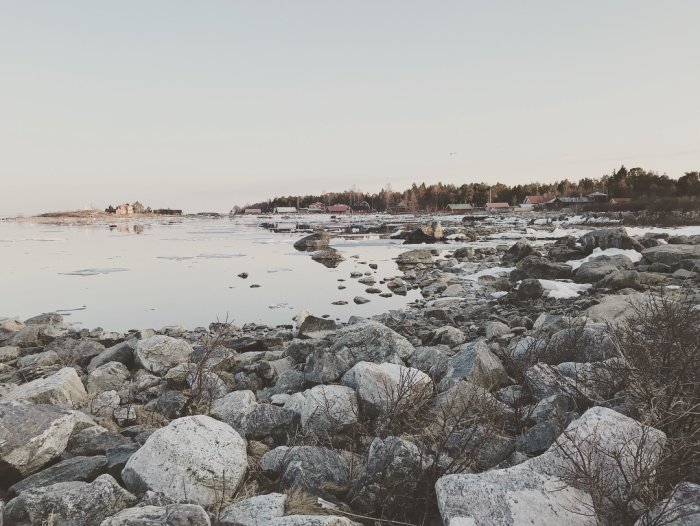Kustlandskap med stenig strand vid skärgård och hus i bakgrunden.