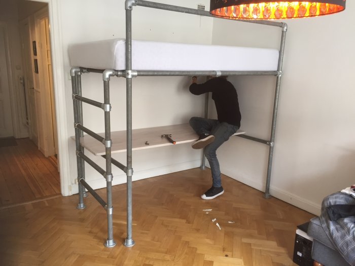 Person bygger loftsäng med integrerad stege och arbetsbord, använder Kee-Clamp kopplingar och rör.