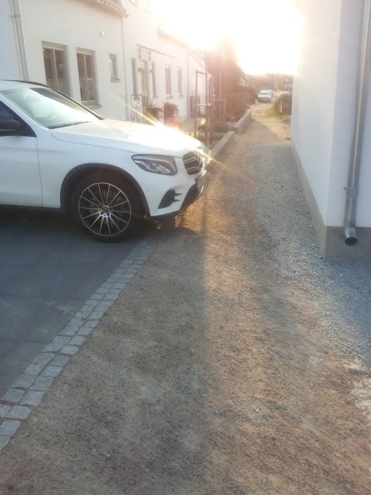En vit bil parkerad i en smal grusdriveway mellan två hus.