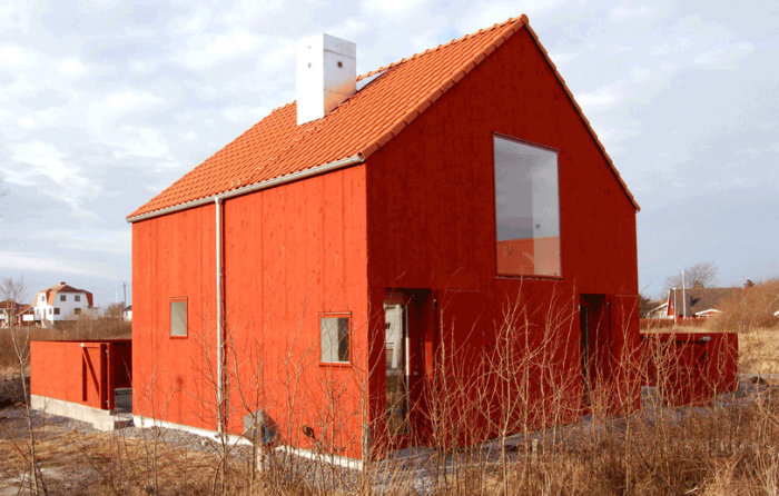 Rödmålat hus med fönster i linje med träfasaden, omgiven av torkade buskar.