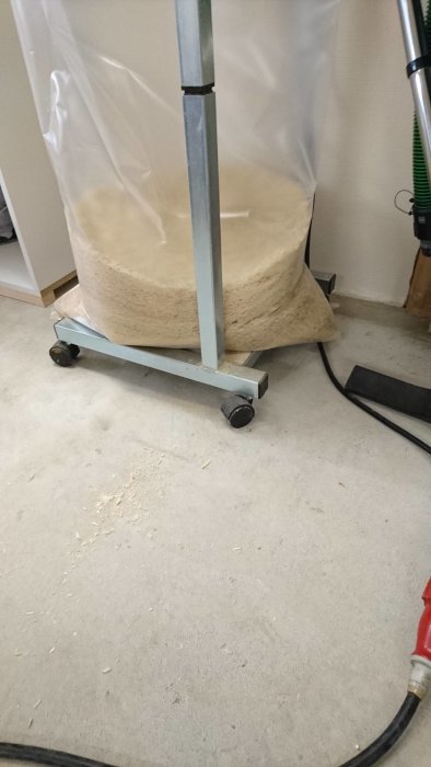 En nästan full, genomskinlig säck på en rullvagn inuti en verkstad, fylld med spån och sågspån på golvet runtomkring.