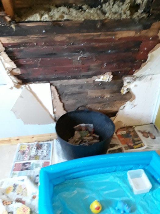 Skadat och öppet tak med synliga träbjälkar och röta, svart hink med spill, tidningar på golvet och blå plastbalja.