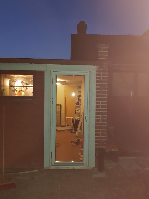 Kvällsbild av ett hus under renovering med öppen dörr, verktyg synliga inne, och en stege vid dörren.