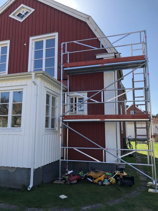 Byggställning uppställd mot en röd husvägg nära fönster och veranda, redo för renoveringsarbete.