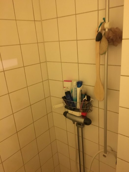 Hörn av ett kaklat badrum med duschstång och hylla med hygienartiklar.