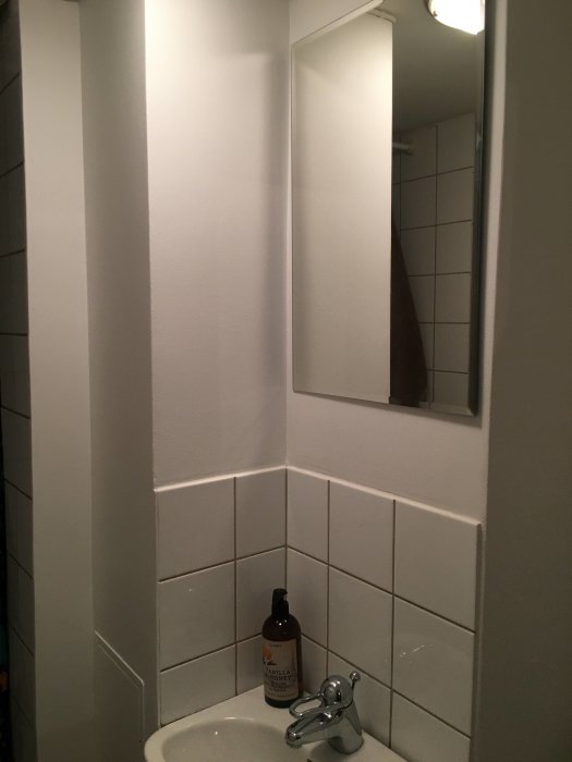 Hörn av ett badrum med vita väggar och handfat, brun tvålflaska och kran, spegel och kakel.