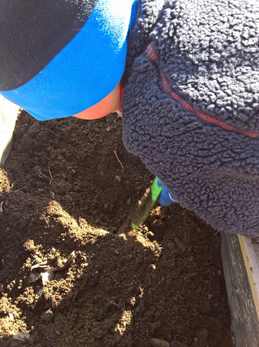 Barn gräver i jordlåda med spade, letar efter något bland mörk jord.