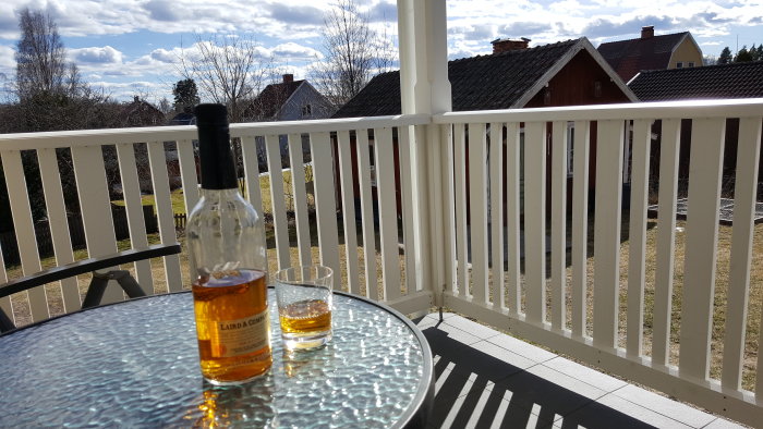 En flaska och ett glas Applejack på ett bord på en veranda med utsikt över hus och träd en solig dag.