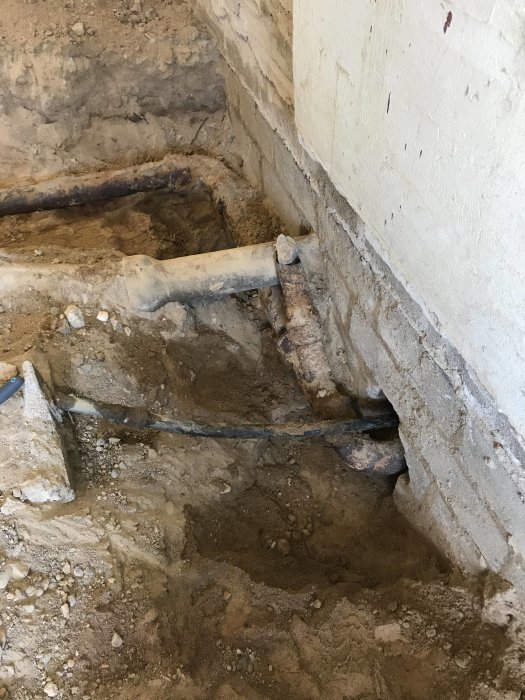 Gammalt gjutjärnsavloppsrör i utgrävd del av betongplatta nära husvägg under renovering.