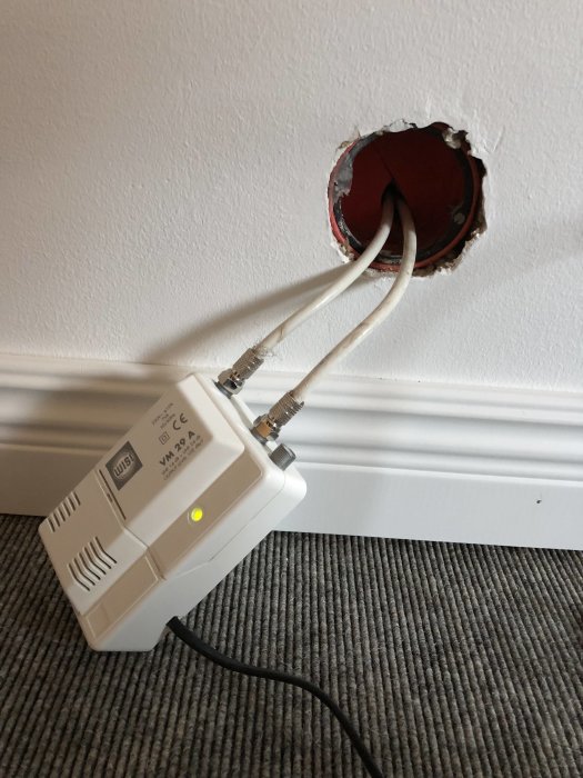 En antennförstärkare med två koaxialkablar anslutna, placerad på ett golv nära en oavslutad väggöppning.