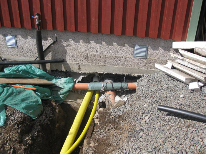 Grävd grop vid husgrund med synligt avloppsrör och byggmaterial runtomkring.