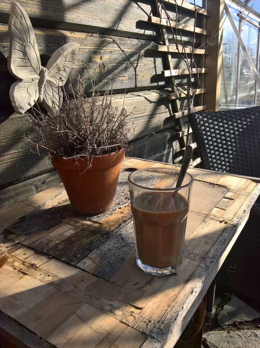 Morgonfika på träbord i pergola med kaffeglas, vissnad planta i kruka och väggdekoration i form av fjäril.