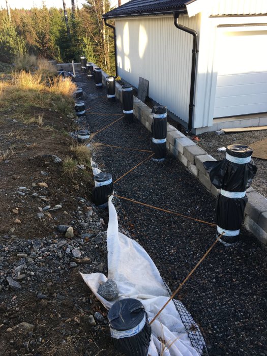 Nyligen färdigställd stenmur längs husvägg med markeringspinnar och förberedelse för gjutning och dränering.