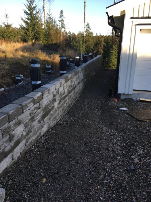 Nybyggd grå mur längs en grusgång med plasttunnor uppställda ovanpå och skog i bakgrunden.