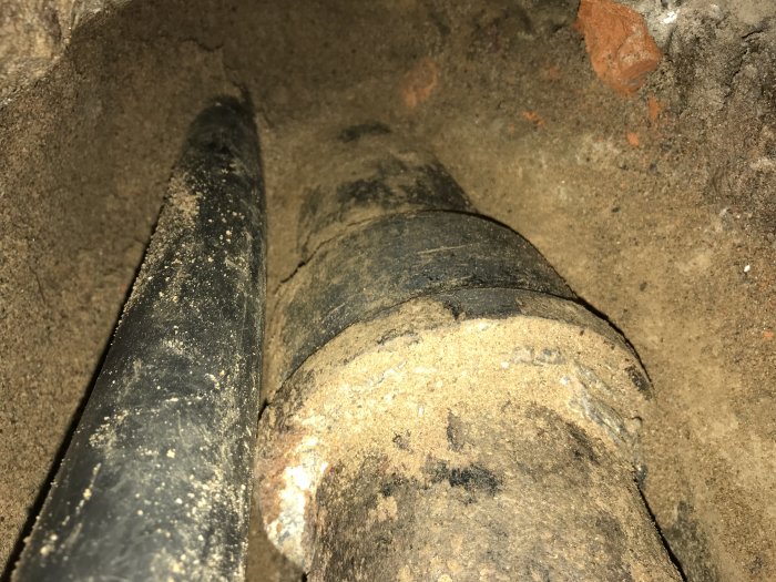 Gammalt gjutjärnsrör i en grävd grop vid en husvägg, indikerar oväntade arkeologiska fynd.