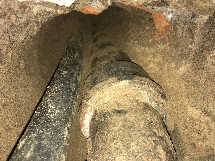 Grävd grop som visar gammalt gjutjärnsrör i marken utanför ett hus.
